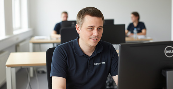 Kevin Zogg, Mitglied des VOLTA Kernteams, sitzt an einem Schreibtisch in einem hellen Büro und arbeitet konzentriert an einem Computer. Er trägt ein dunkelblaues Poloshirt mit Karakun-Logo. Im Hintergrund sind zwei weitere Mitarbeitende an ihren Arbeitsplätzen unscharf zu erkennen. Tageslicht fällt von links durch große Fenster und sorgt für eine natürliche, freundliche Ausleuchtung der Szene.